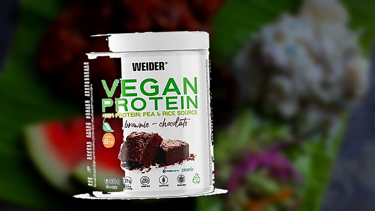 Bote de proteína vegana de chocolate Weider sobre mostrador de mármol, con scoop de polvo, nibs de cacao y shaker, escena de cocina-gimnasio matutina con iluminación suave.