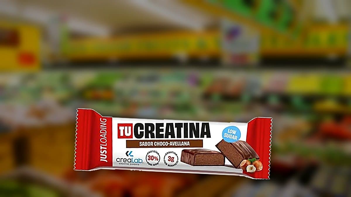Barrita de creatina Just Loading sabor chocolate-avellana sostenida en mano cerca de bolsa de gym, con etiqueta nutricional parcialmente visible bajo luz natural.
