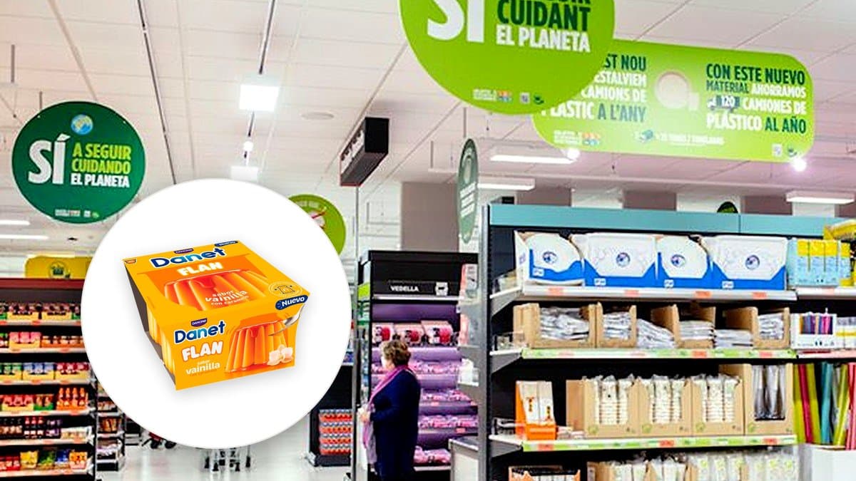 Estantería refrigerada de supermercado llena de yogures, con tubs de marca blanca a un lado y packs de Danone al otro, luz fría de refrigeración, cliente desenfocado en el pasillo