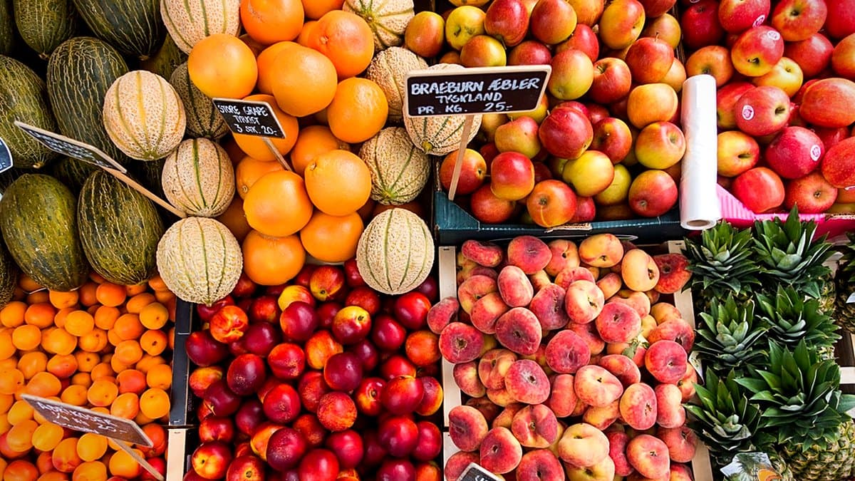 Aisle de frescos en supermercado con tomates en cajas, botellas de aceite de oliva al fondo, iluminación natural cálida