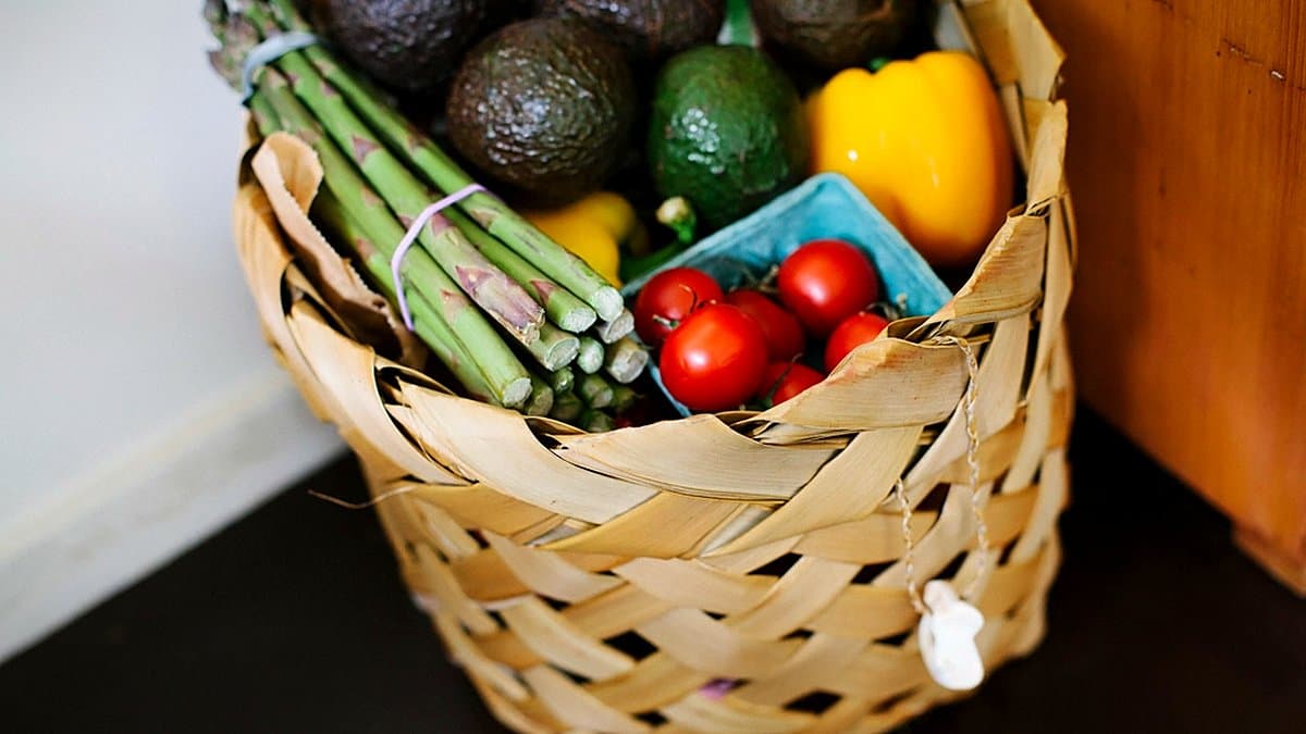 Cesta semanal de compra sobre mesa de madera con tomates, calabacín, huevos, conservas, lechuga y lista manuscrita de compra, luz natural cenital.