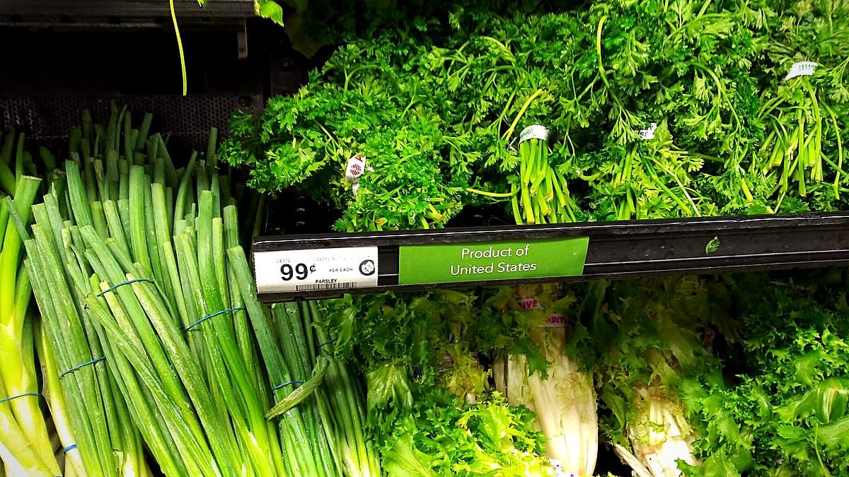Caja de supermercado con cliente al fondo colocando productos sobre la cinta transportadora, personal de caja enfocado, luz natural de tienda, sin textos superpuestos