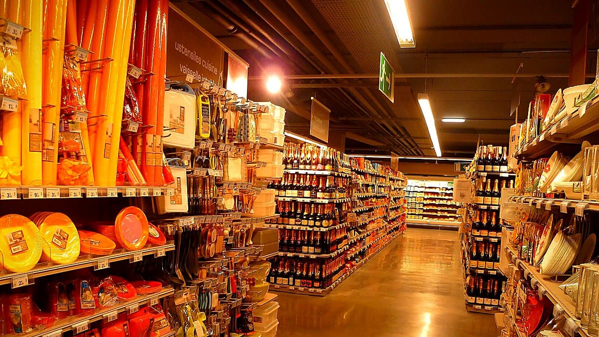 Carrito de compra lleno en pasillo de supermercado, comprador revisando precios con expresión preocupada, iluminación tienda